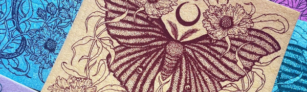 A yellow Swedish Dishcloth printed with Fennec Design's moth and botanical print lays on top of other multi-colored cloths