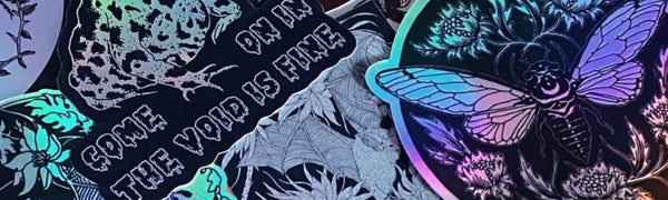 A multitude of black and silver holographic stickers from Fennec Design lay glimmering. One reads "COME ON IN, THE VOID IS FINE". The designs feature toads, moths, bats, and other natural elements.
